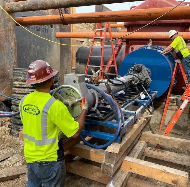 Two workers wearing helmets standing next to a large blue pipeline with a wheel, placed on a wooden structure.