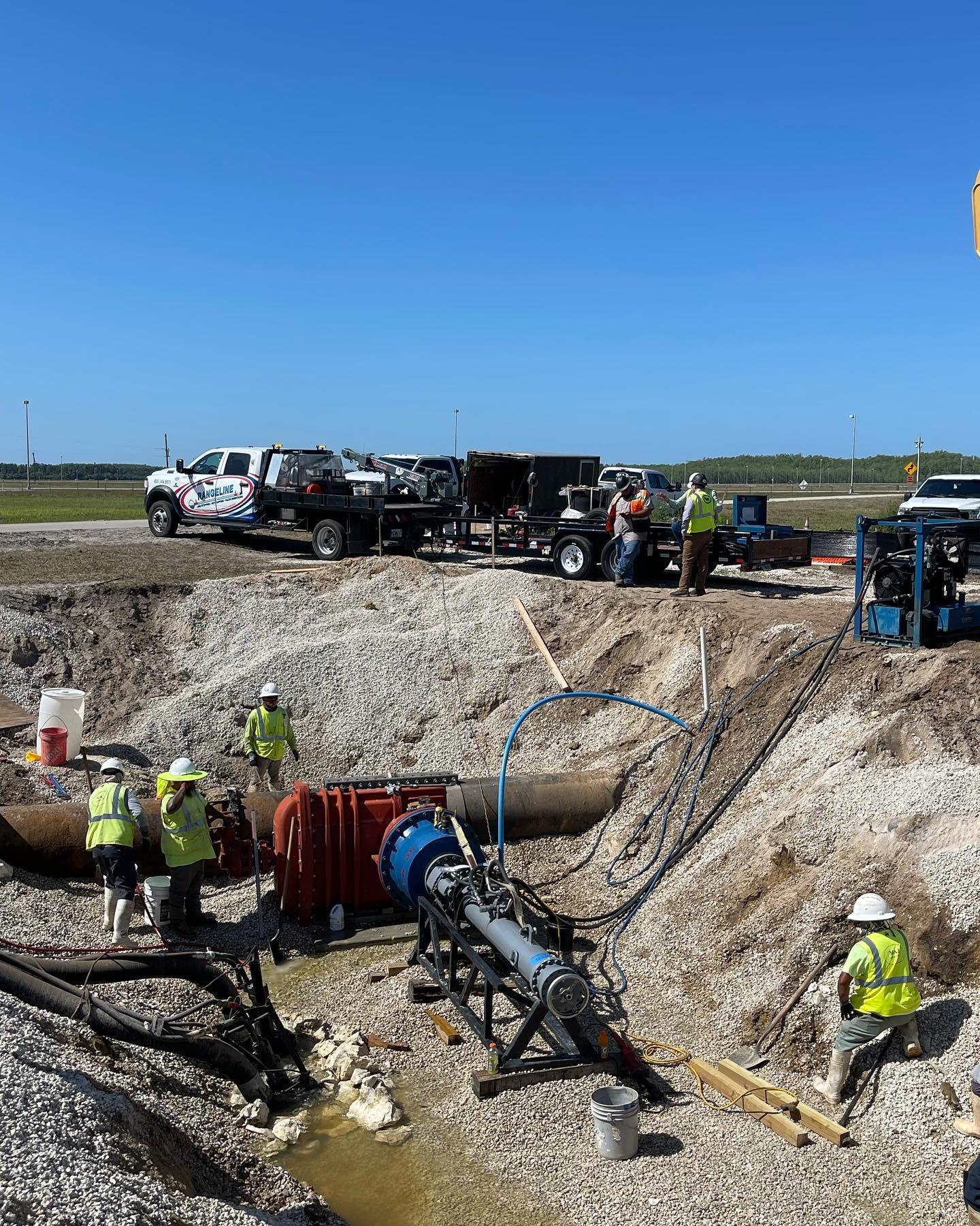 A large hole in a deserted area with a large pipe coming out in the middle with multiple workers around it.