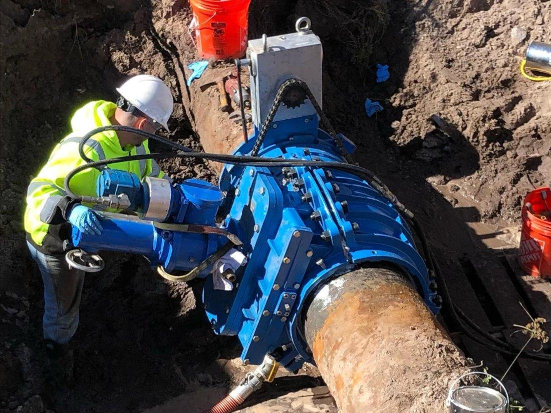 Utility worker servicing a large blue pipeline valve assembly inside a trench during underground water infrastructure maintenance.