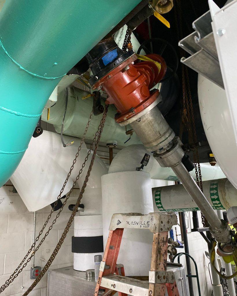 A red pipe tool attached beneath a large green pipe, with chains and a ladder in an industrial utility room.