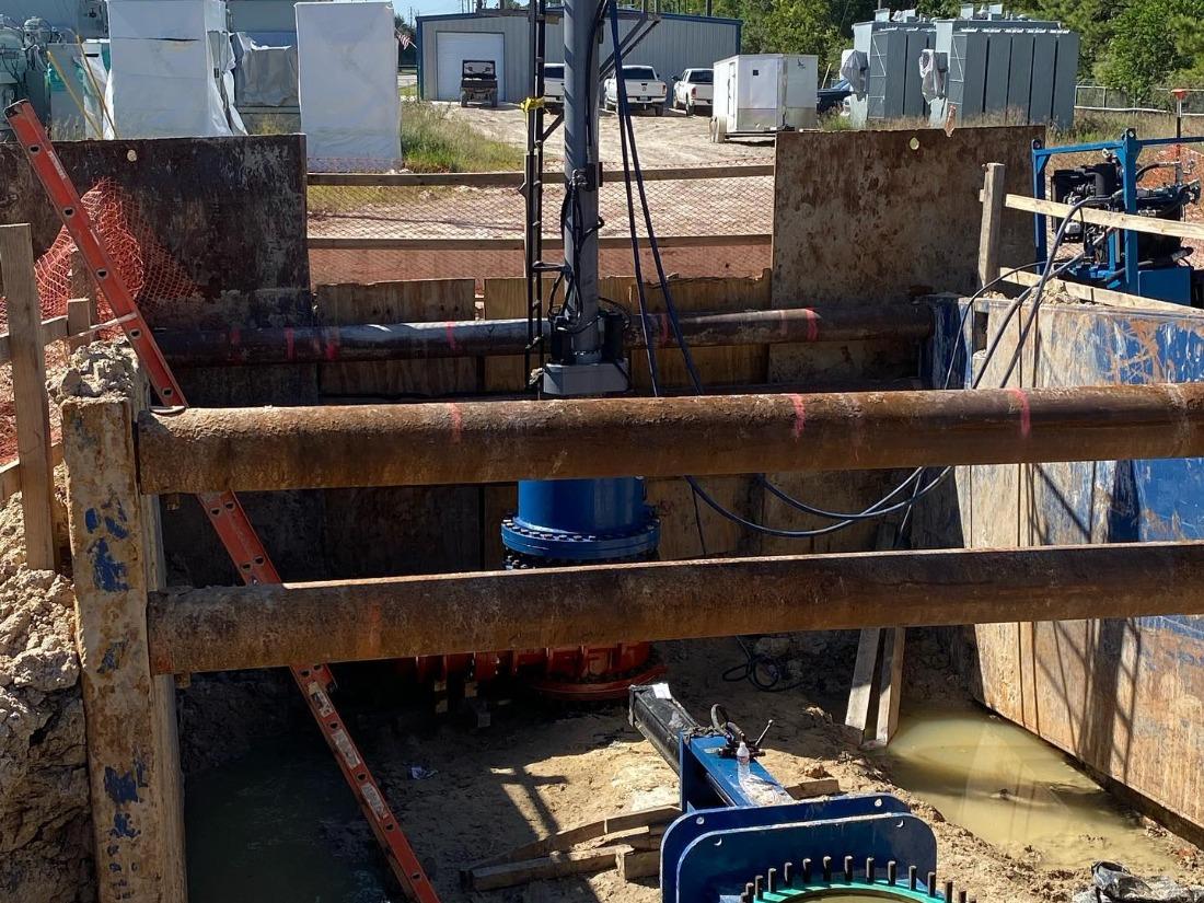 A valve assembly rises from a muddy excavation, with metal shoring around it and work equipment beyond the pit.