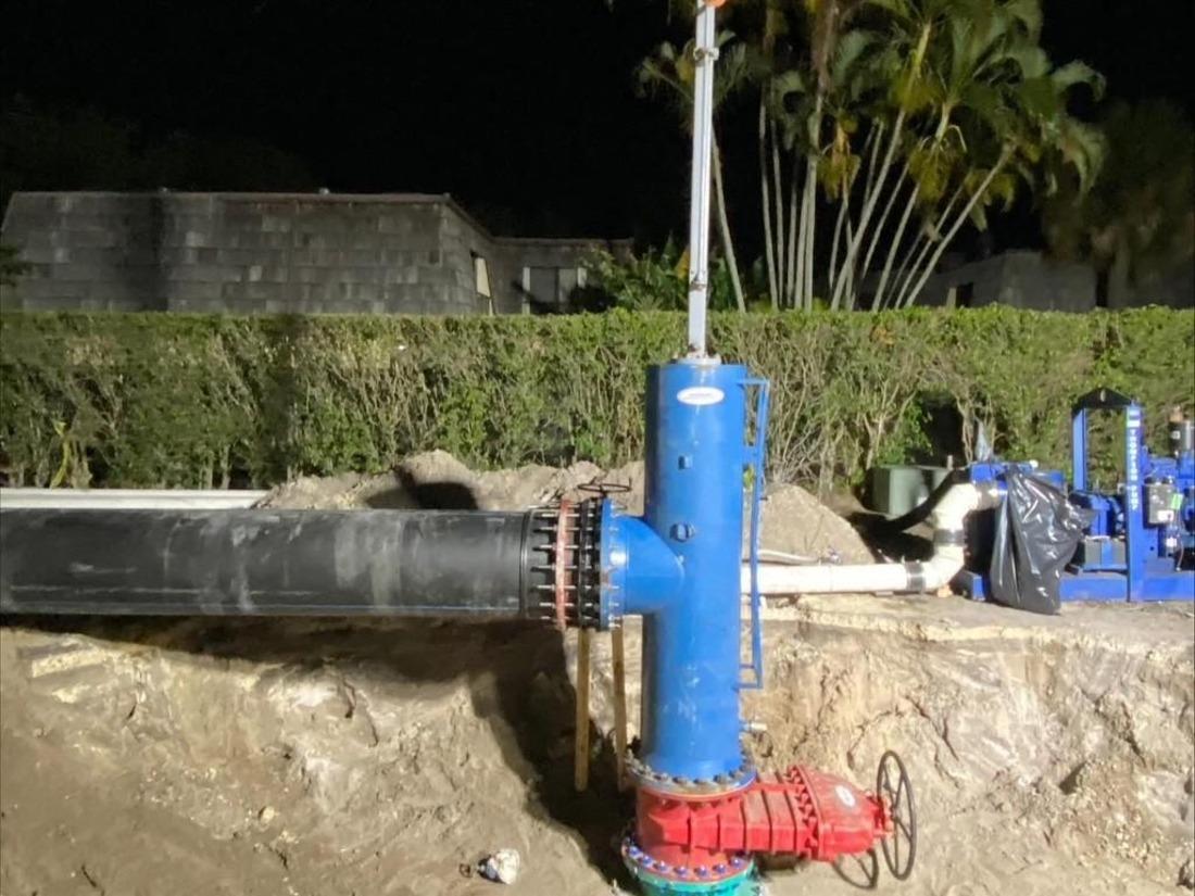 A trench that contains a blue pipe column and a red valve connected to black piping near hedges and palms.