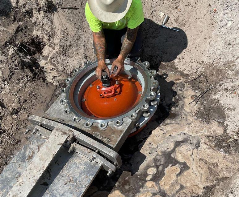 A worker wearing a yellow shirt and a white hat touching the top bolt of a metal valve placed close to the ground.