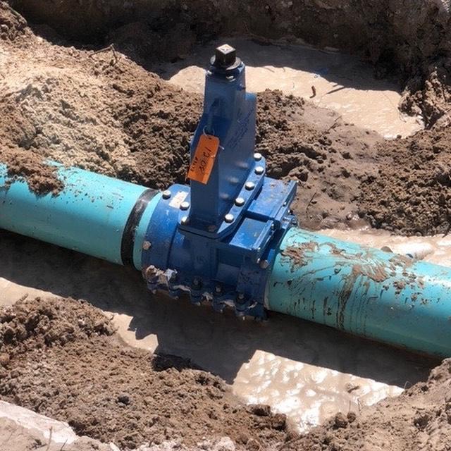 A thin blue pipe underground with an exposed metal valve attached to the top, sealed with a large metal bolt.