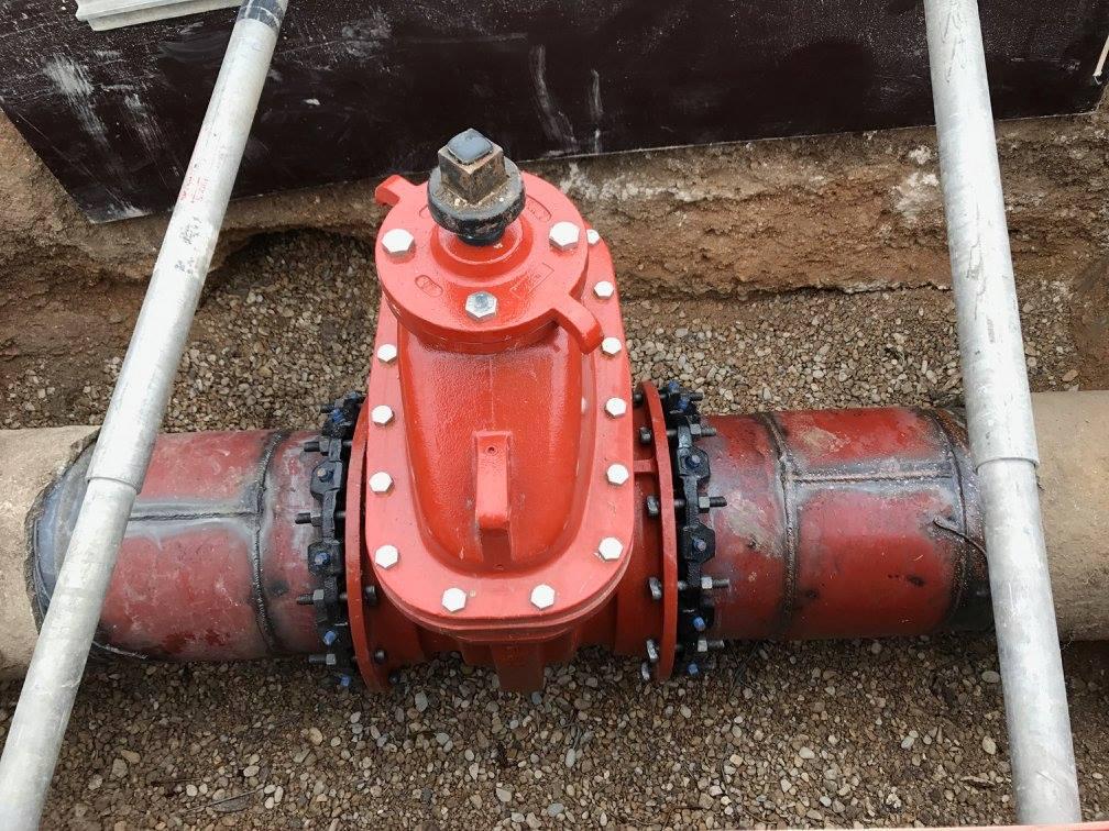 A close-up of a connecting point on a pipe painted with a red coating, secured with multiple bolts placed on dirt.